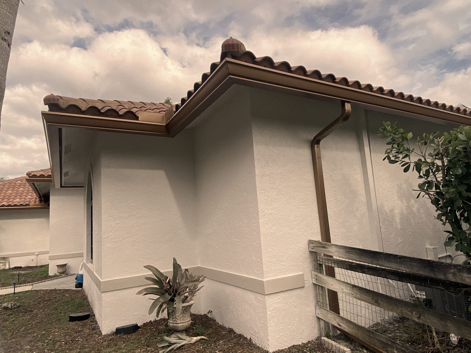 A bronze gutter and downspout follow the eave of a white stucco house with a barrel tile roof.