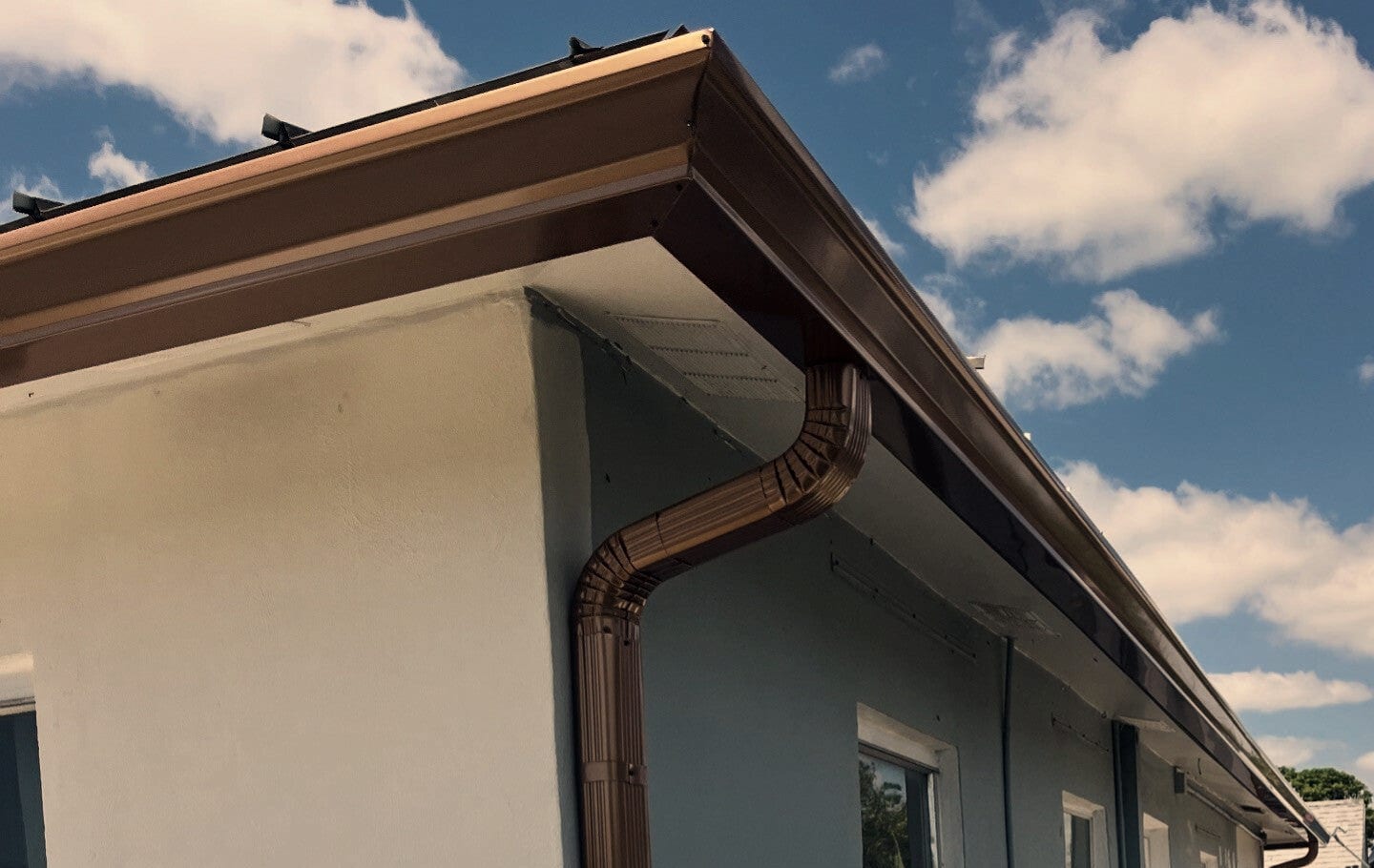 A copper-tone seamless gutter turns the corner of a pale green stucco building and feeds a matching downspout.
