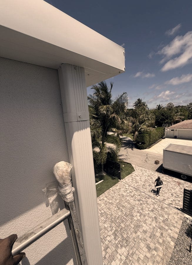 Seen from the top of a ladder, a new white gutter and downspout run along the eave of a gray stucco home while a crew member carries the next section across the paver driveway below.