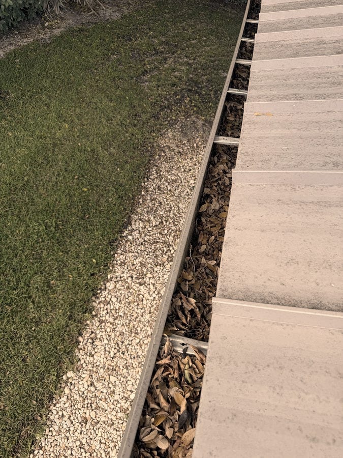A gutter run packed end to end with dried leaves, photographed from the roof above a gravel bed and lawn.