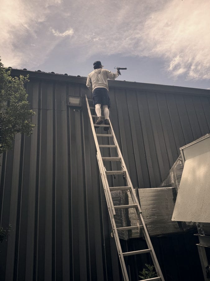 A technician on an extension ladder seals a gutter joint with a caulking gun along the roofline of a dark metal commercial building.