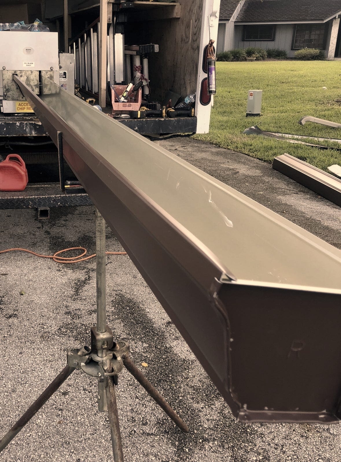A brown seamless gutter is formed to length on site, running out of the truck-mounted machine onto a roller stand in the driveway.